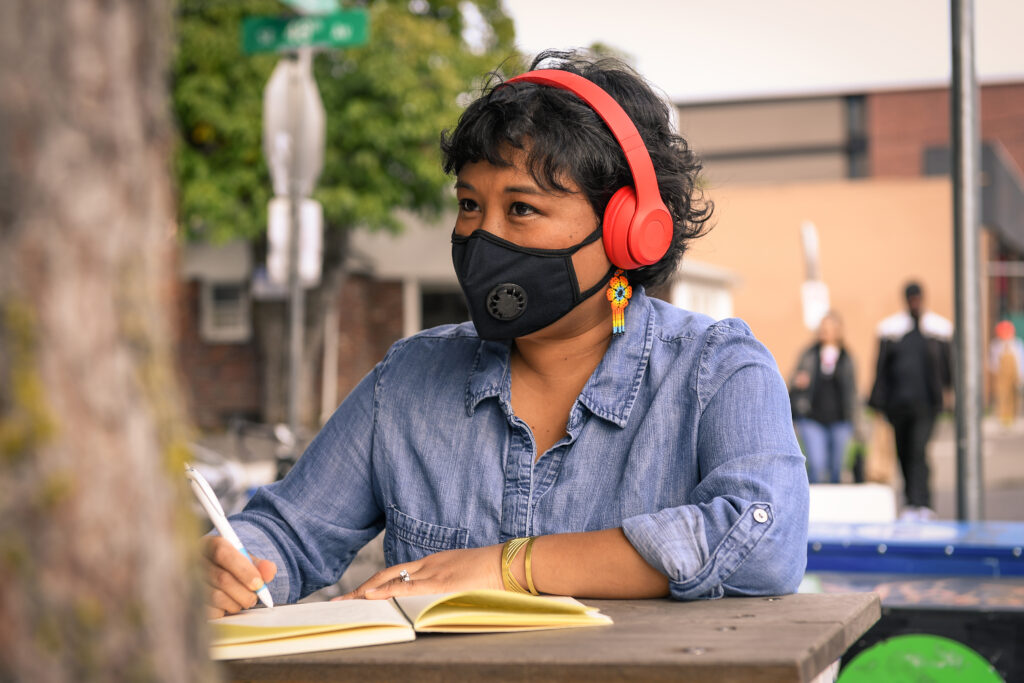 Close-up of a woman with a filtering face mask, sitting at a table with notebook and pen. She has colorful flower earrings and headphones on while looking into the distance.
