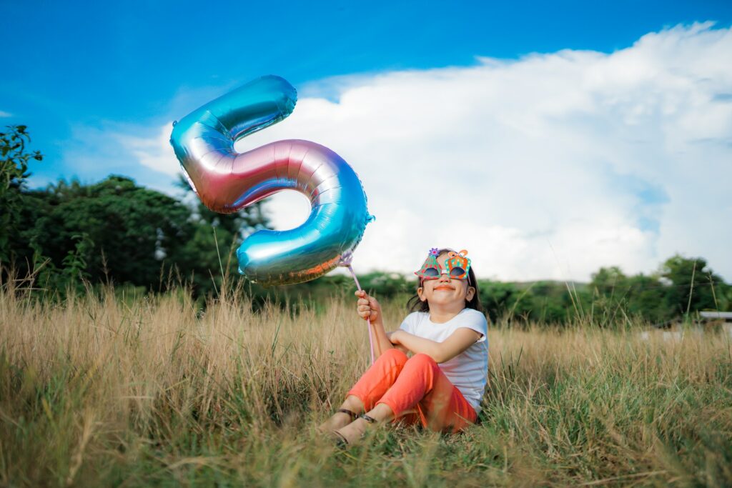 A girl with zany sunglasses sitting in a field and holding a balloon shaped like the number five.